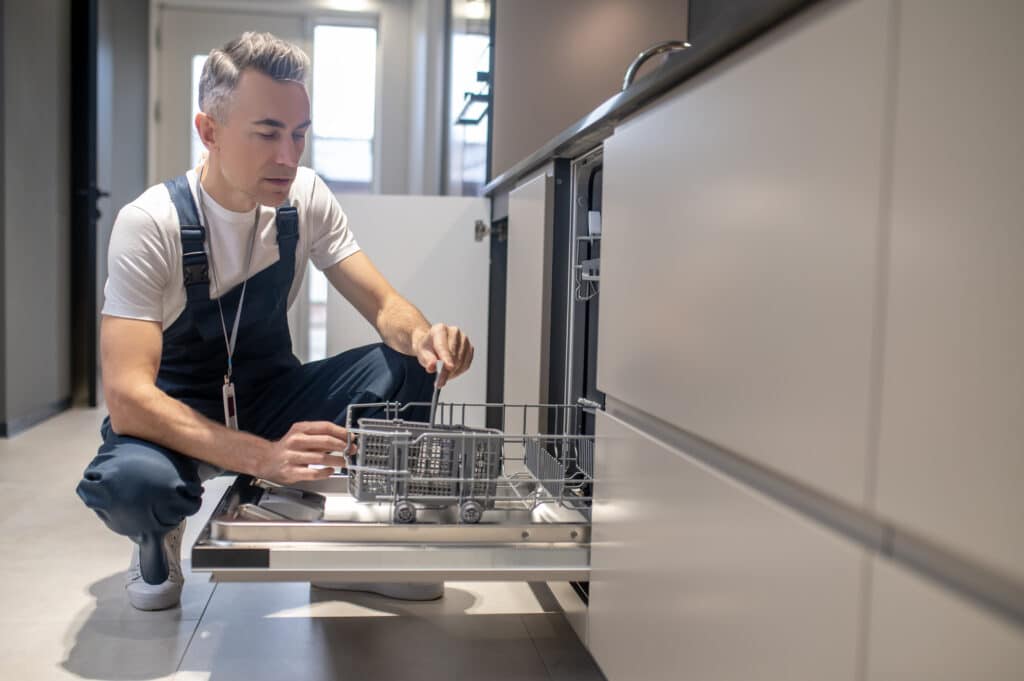 Dishwasher inspection. Involved attentive man in white tshirt and blue overalls with badge crouching down opening dishwasher looking in modern kitchen during day